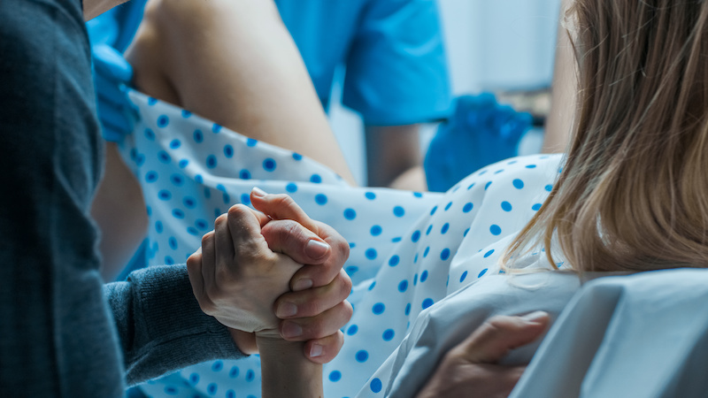 Emergency In the Hospital: Woman Giving Birth, Husband Holds Her Hand in Support, Obstetricians Assisting. Modern Delivery Ward with Professional Midwives; Blog: Stages of Labor Explained