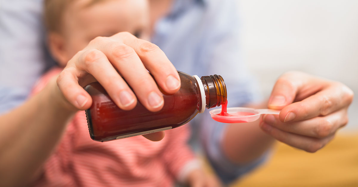 close up of mother pouring cough syrup. blurred baby boy in background; blog: Commonly Prescribed Pediatric Medications